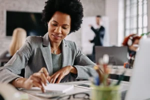smiling-black-entrepreneur-working-financial-reports-her-desk-office-there-are-people-background-scaled