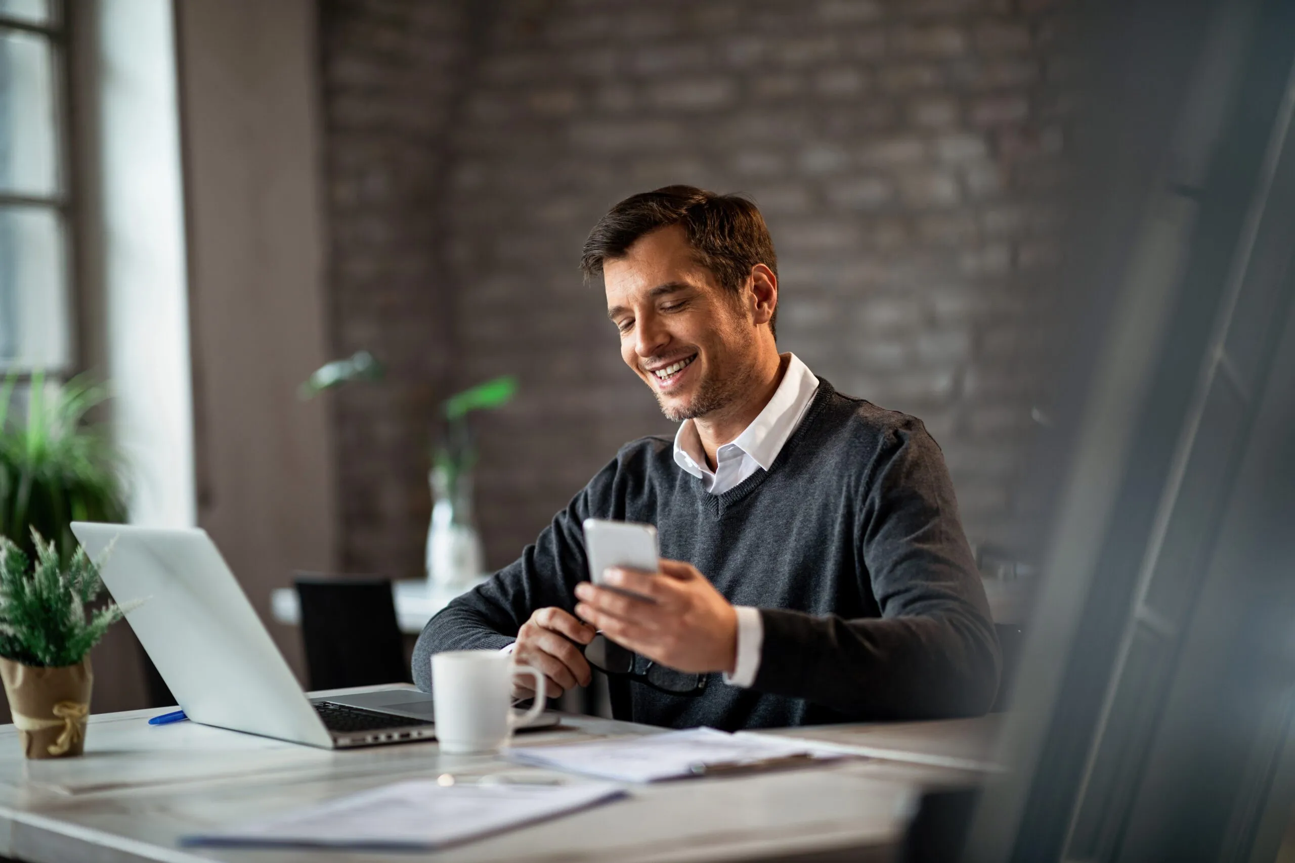 happy-businessman-texting-mobile-phone-while-working-laptop-office-scaled
